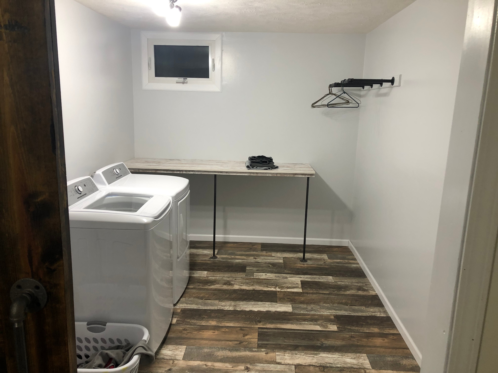 Finished basement laundry room with a wood-look folding counter on black pipe legs, a wall-mounted hanging rack, and plank-style vinyl flooring
