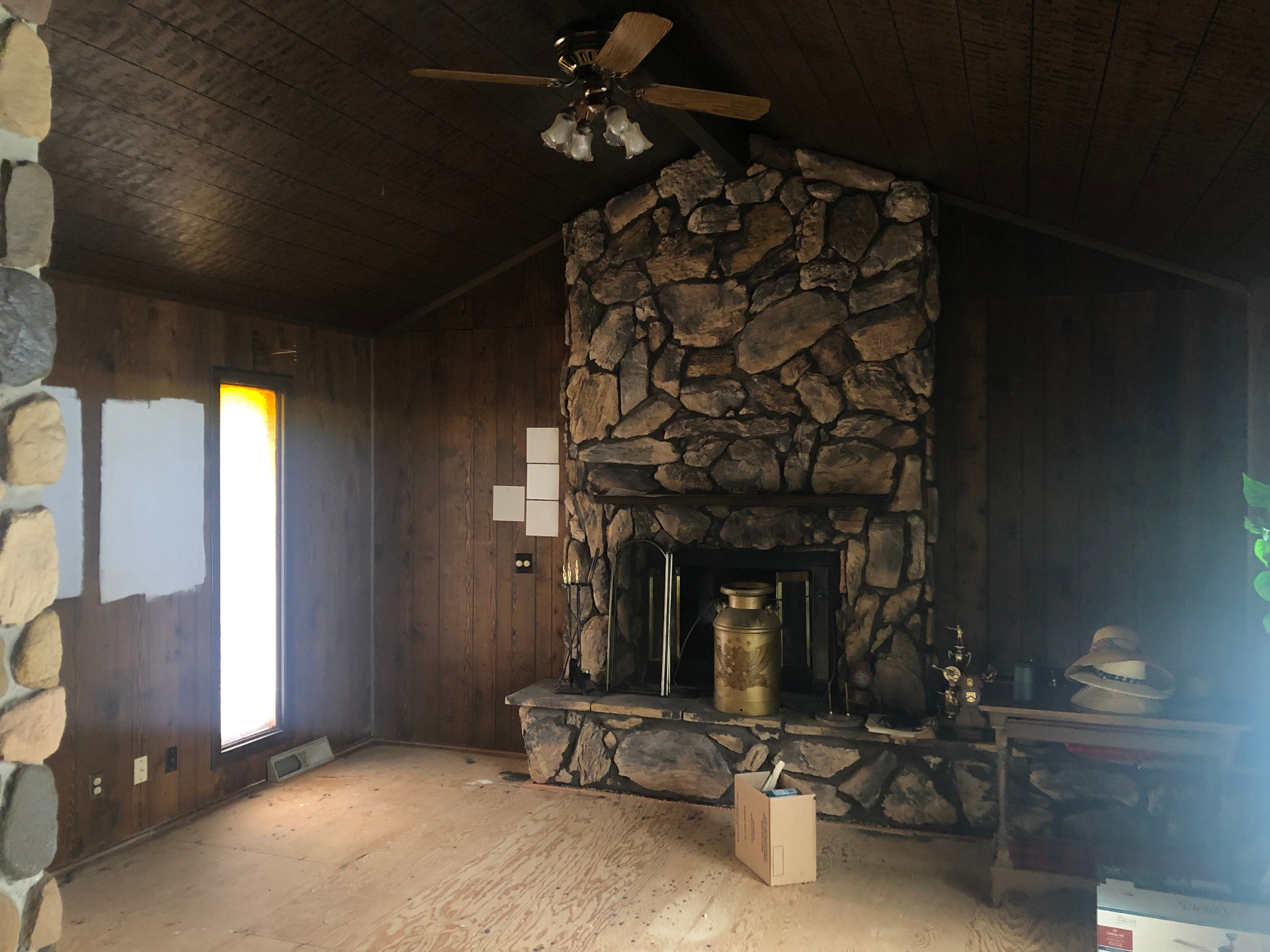 Cleared living room with dark wood plank ceiling, dark wood paneling, and a large floor-to-ceiling stone fireplace, with paint test squares on the wall