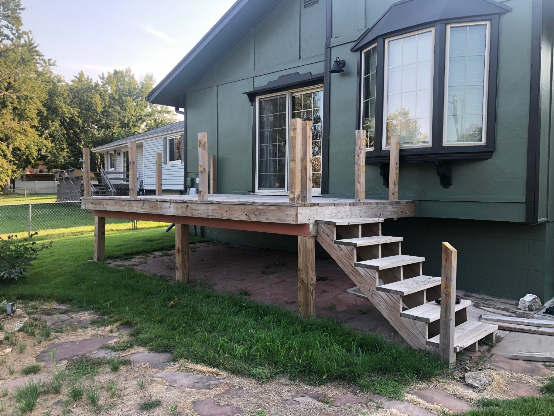 Deck framed and decked but not yet stained, with vertical railing posts and open stair stringers running down to a paver patio