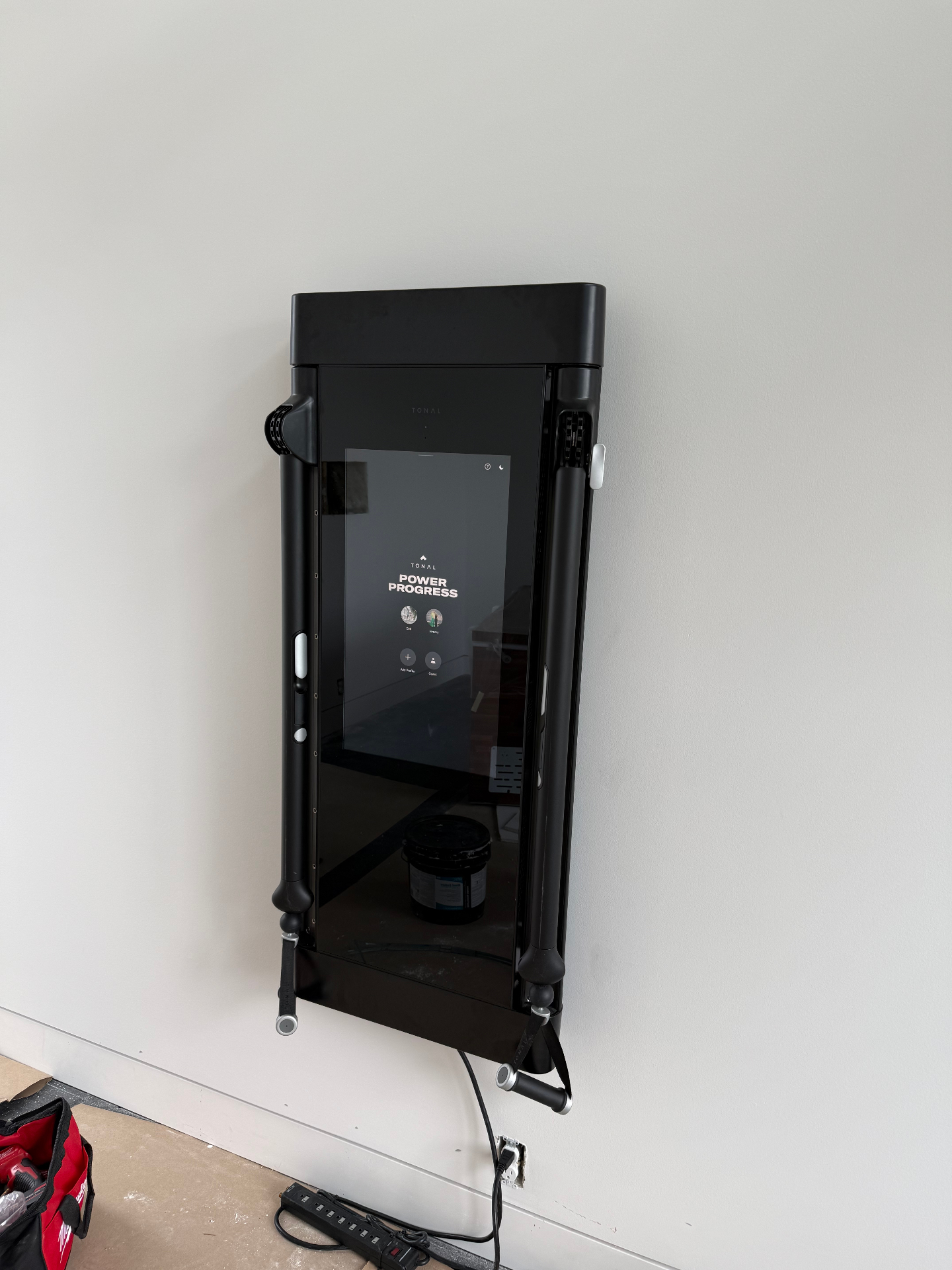 Angled detail of a Tonal wall-mounted smart home gym installed flush and level on a white wall, powered on and showing the profile screen