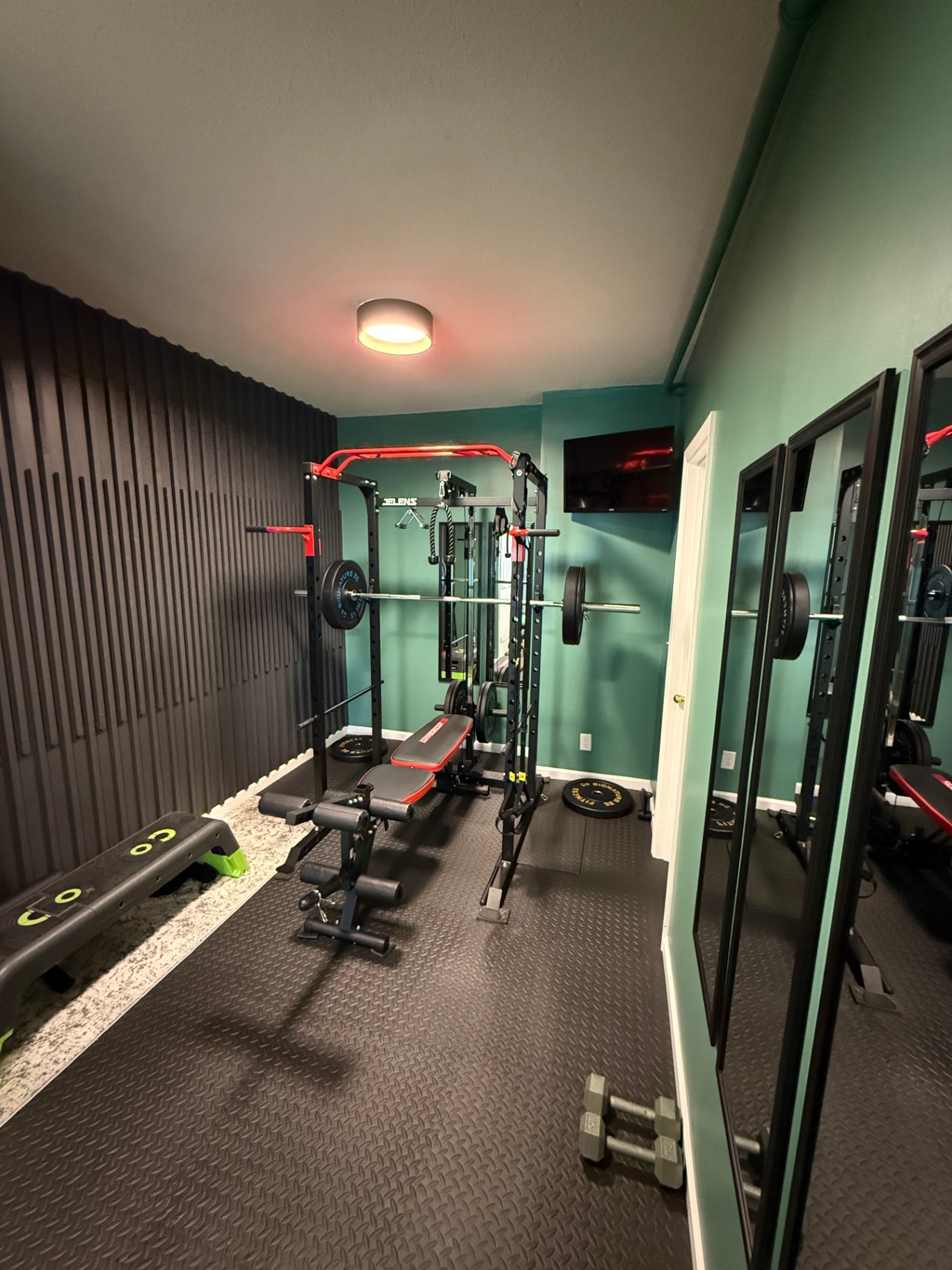 Finished home gym with a vertical black slat accent wall, teal painted walls, black rubber tile flooring, power rack with bench, wall-mounted TV, and full-length mirrors