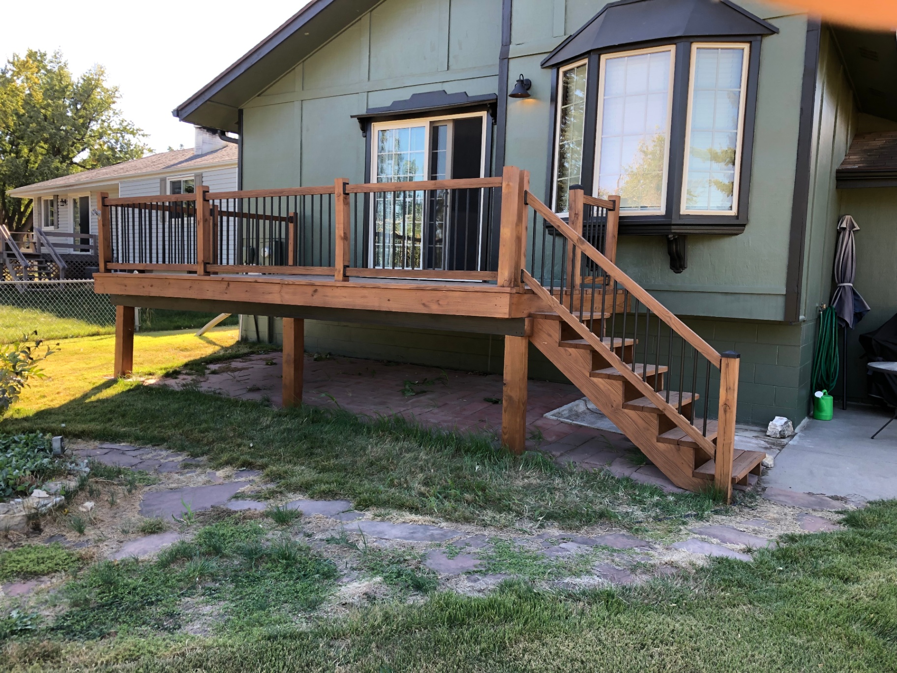 Finished cedar-toned deck attached to a green house, with wood railings, black metal balusters, and a stained stair run down to a paver patio