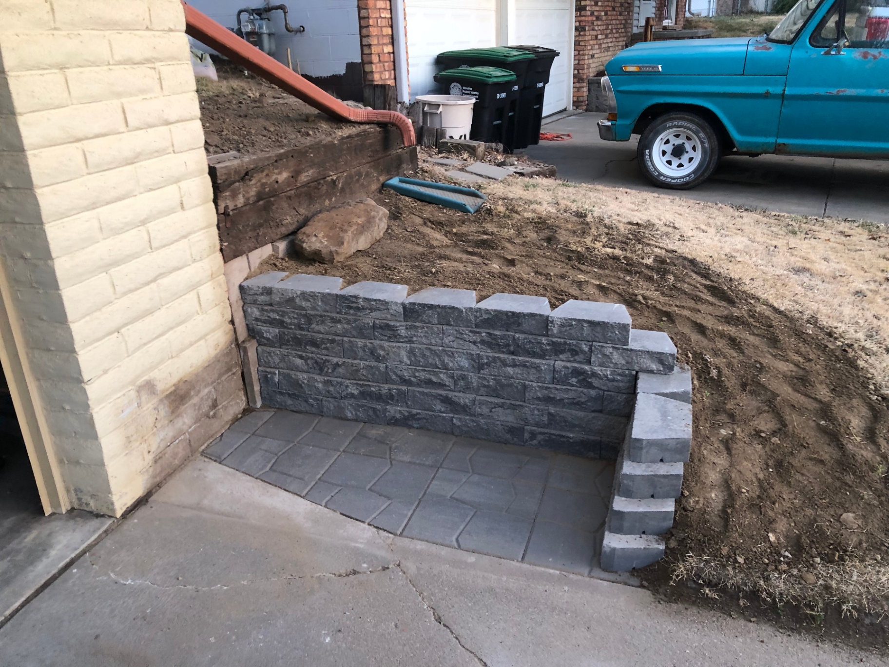 Finished stacked-block retaining wall and paver pad tucked against a painted brick garage wall, ready for trash bins to sit level against the house