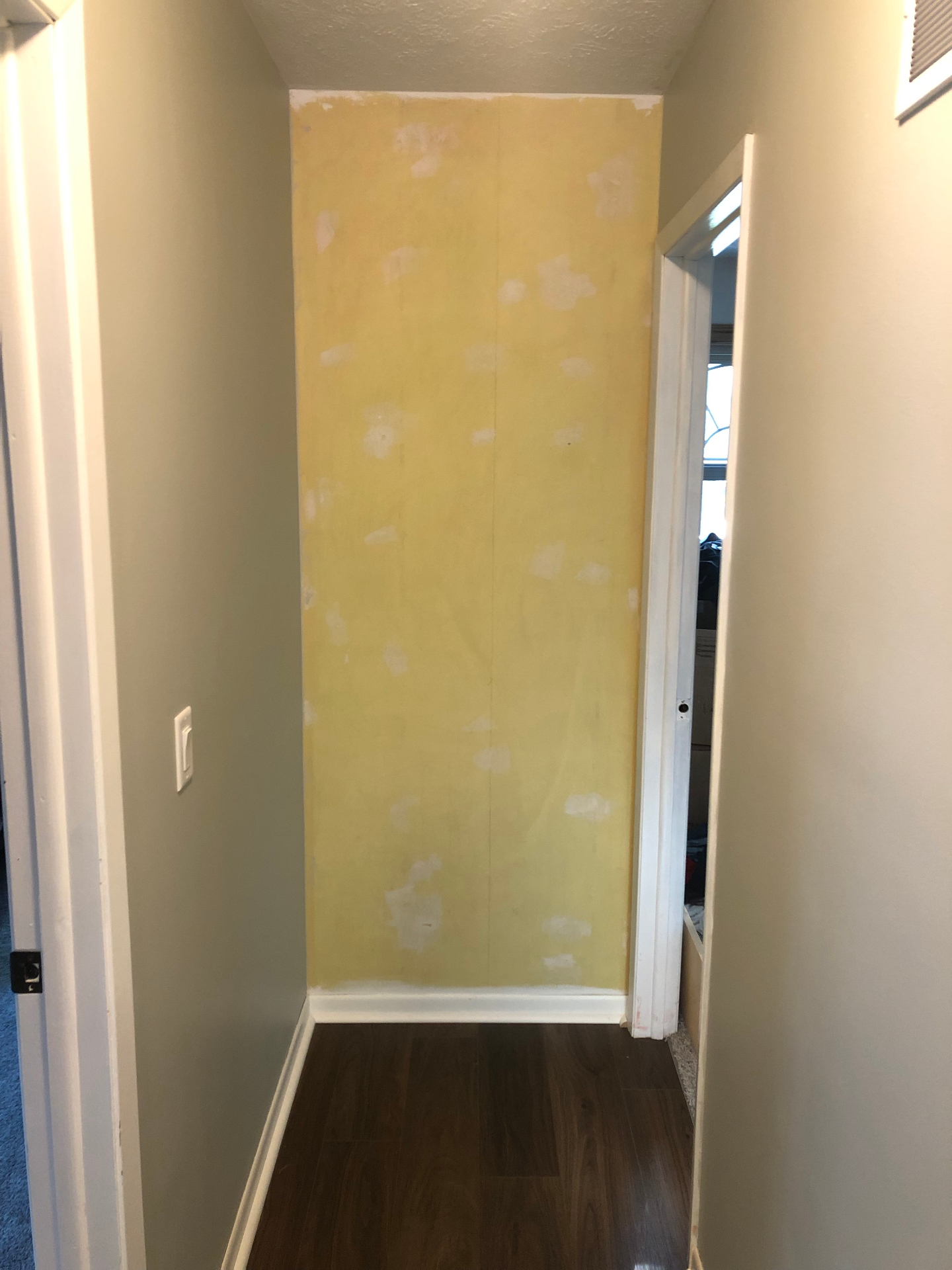 Narrow hallway end wall in original yellow paint with visible patch spots, next to grey side walls and dark wood-look flooring