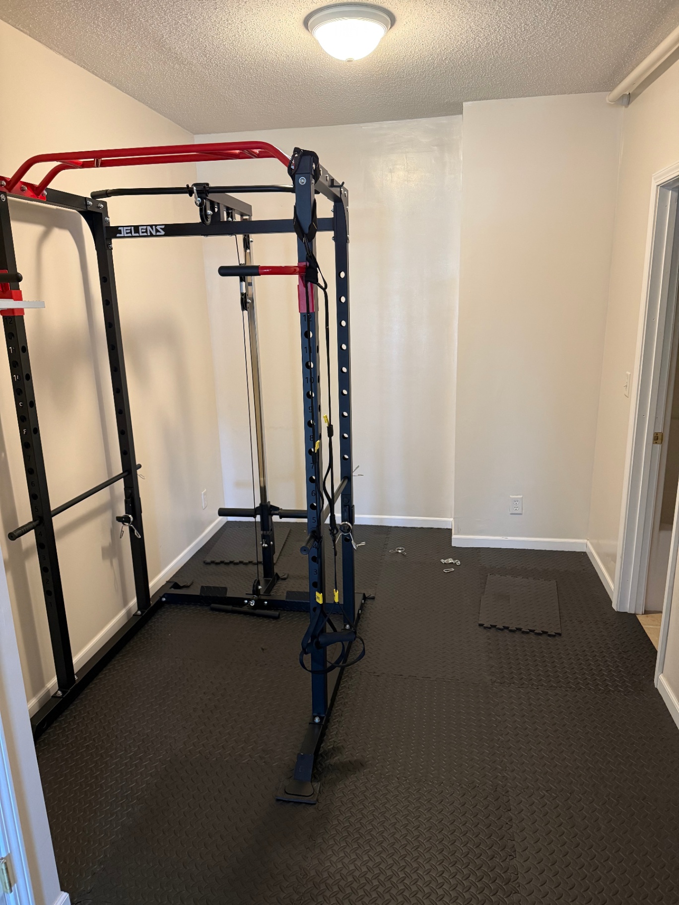 Small workout room with plain white walls and a power rack sitting on interlocking rubber floor tiles before any finish work