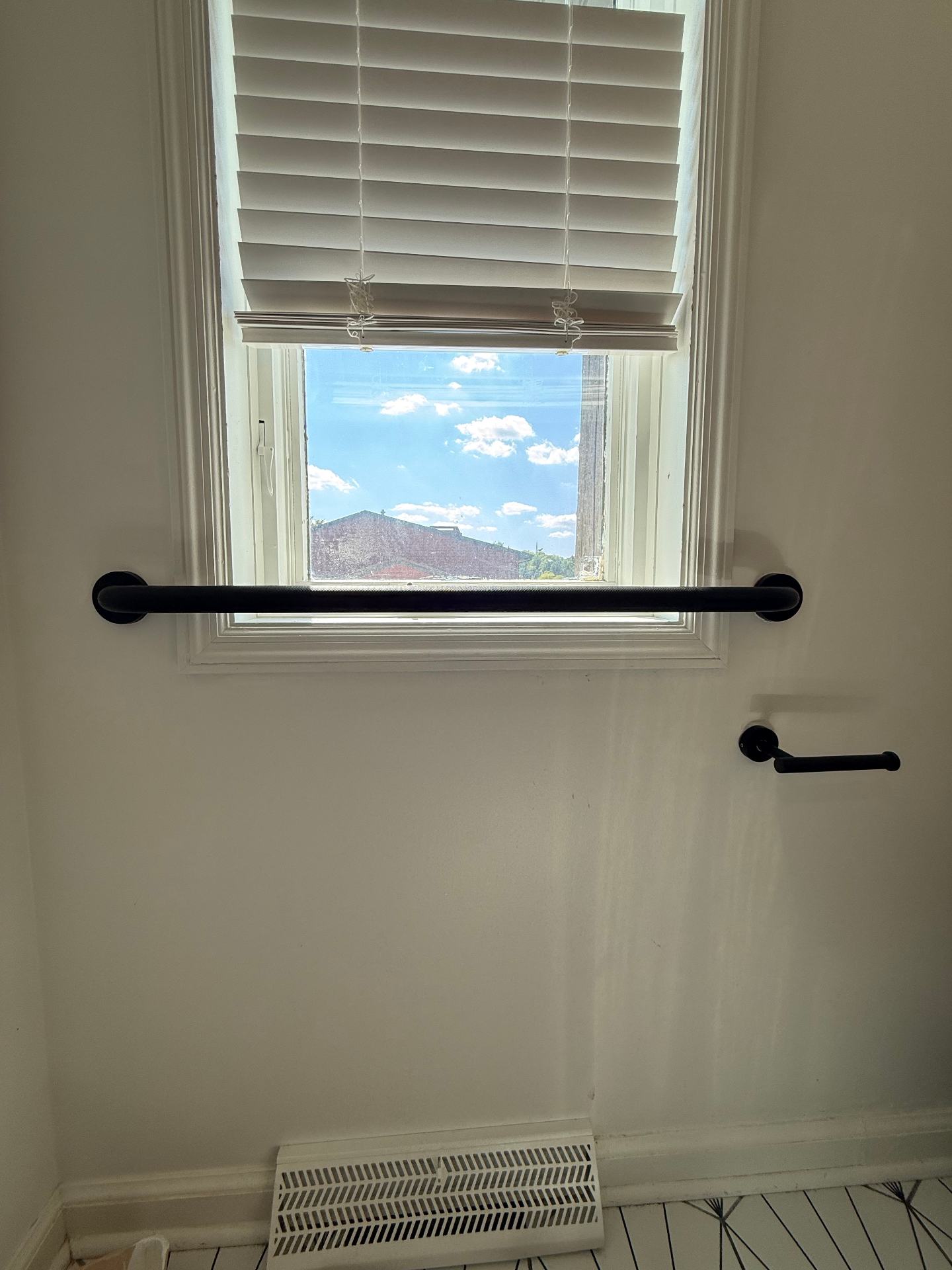 Matte black bathroom grab bar mounted horizontally under a window with a matching matte black toilet paper holder on the adjacent wall