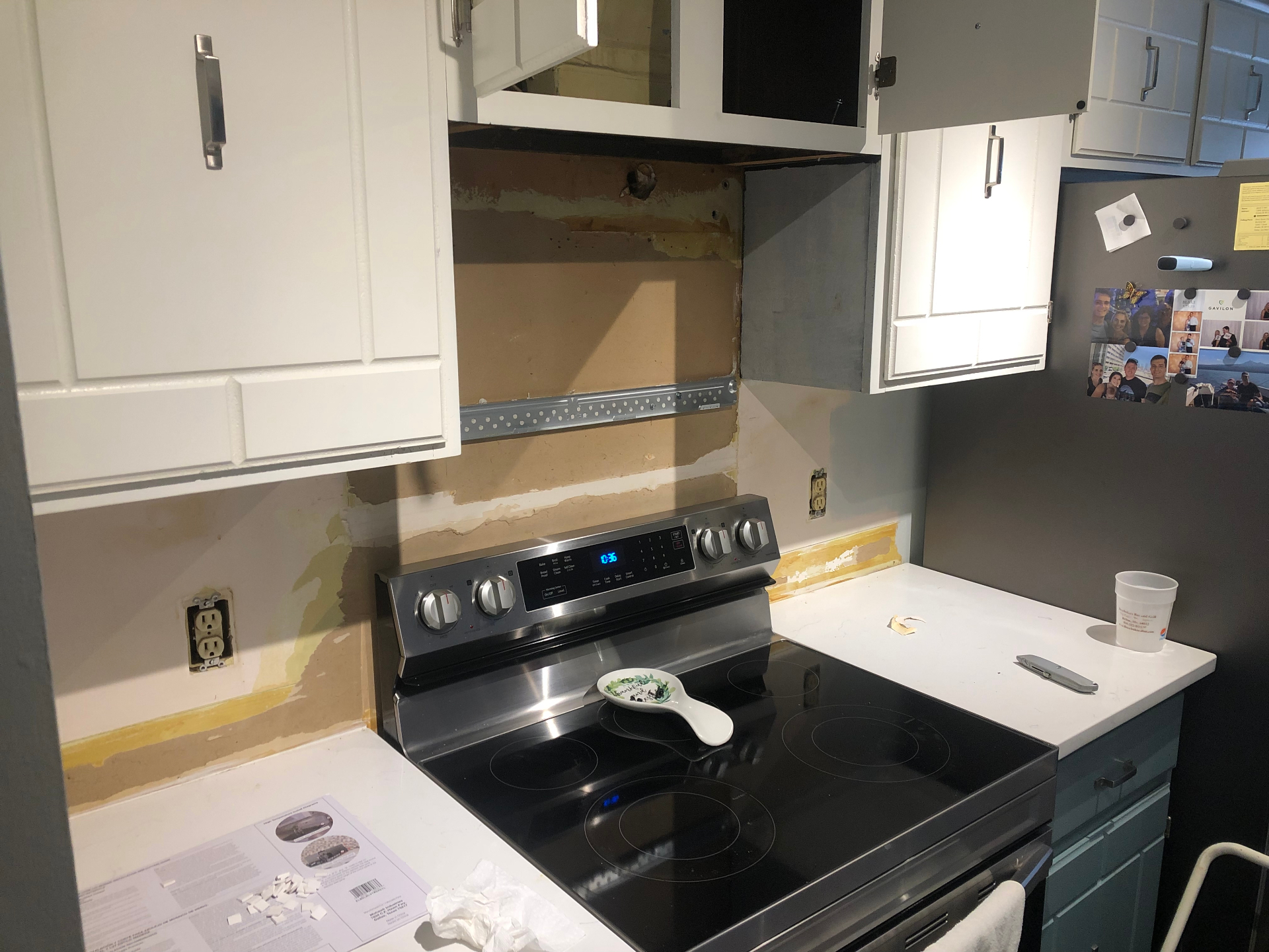 Kitchen wall with existing backsplash removed, exposing patched drywall and mounting points behind the range between white cabinets