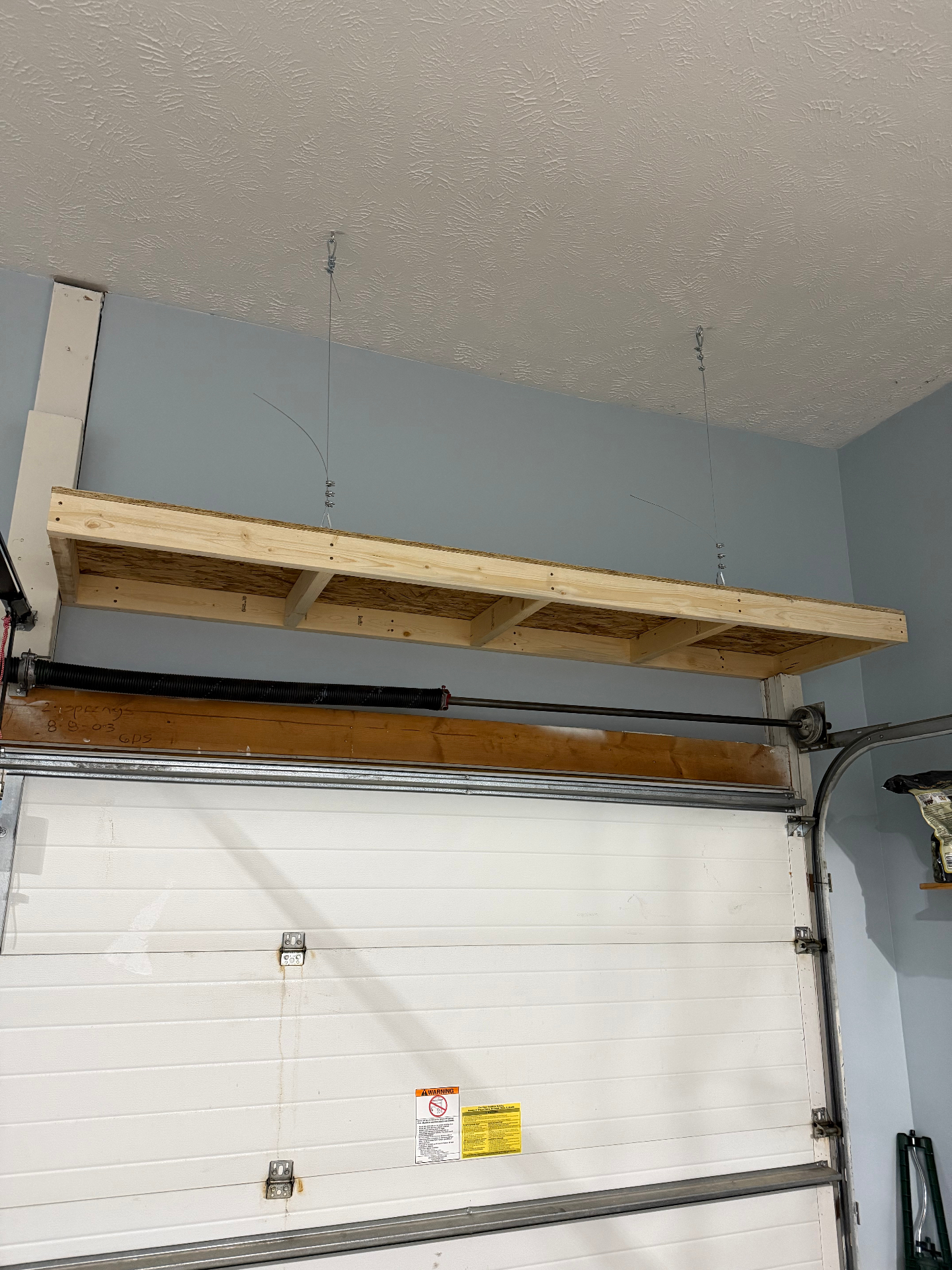 Freshly built wood overhead storage shelf suspended from the garage ceiling above the garage door with steel cables and hooks