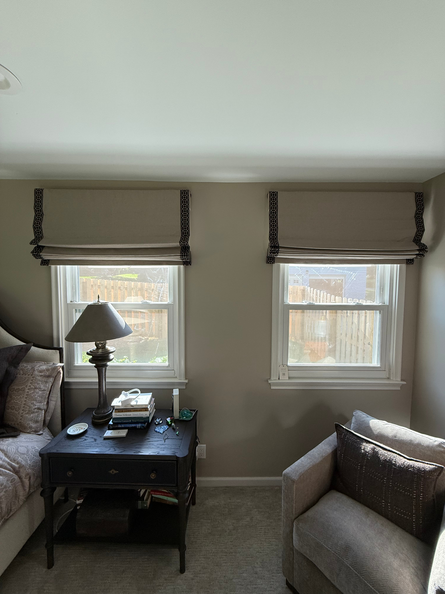 Two windows in a bedroom fitted with linen roman shades trimmed with a dark geometric border