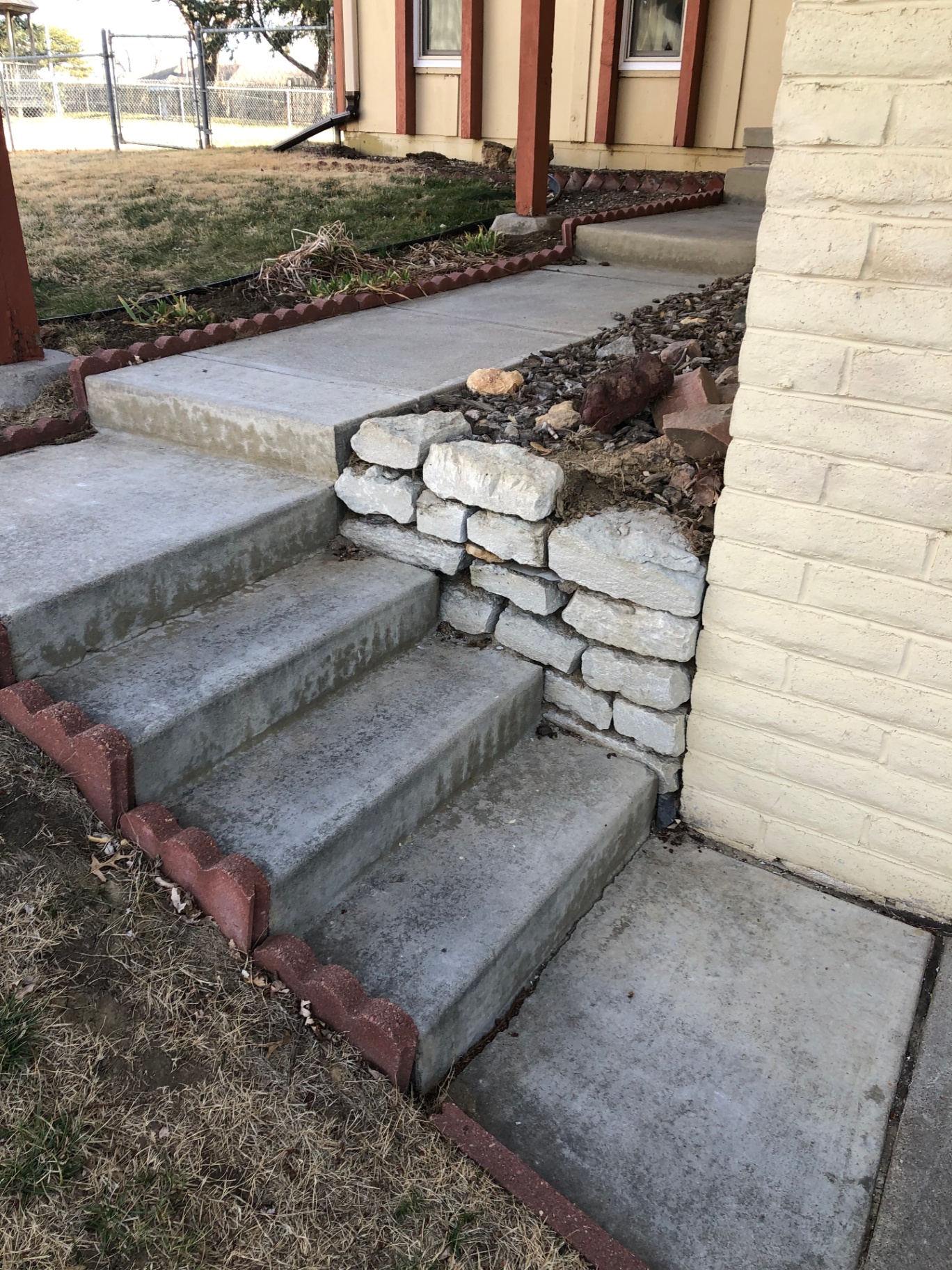 Front concrete steps next to a loose, crumbling stacked-stone retaining wall along a painted brick foundation