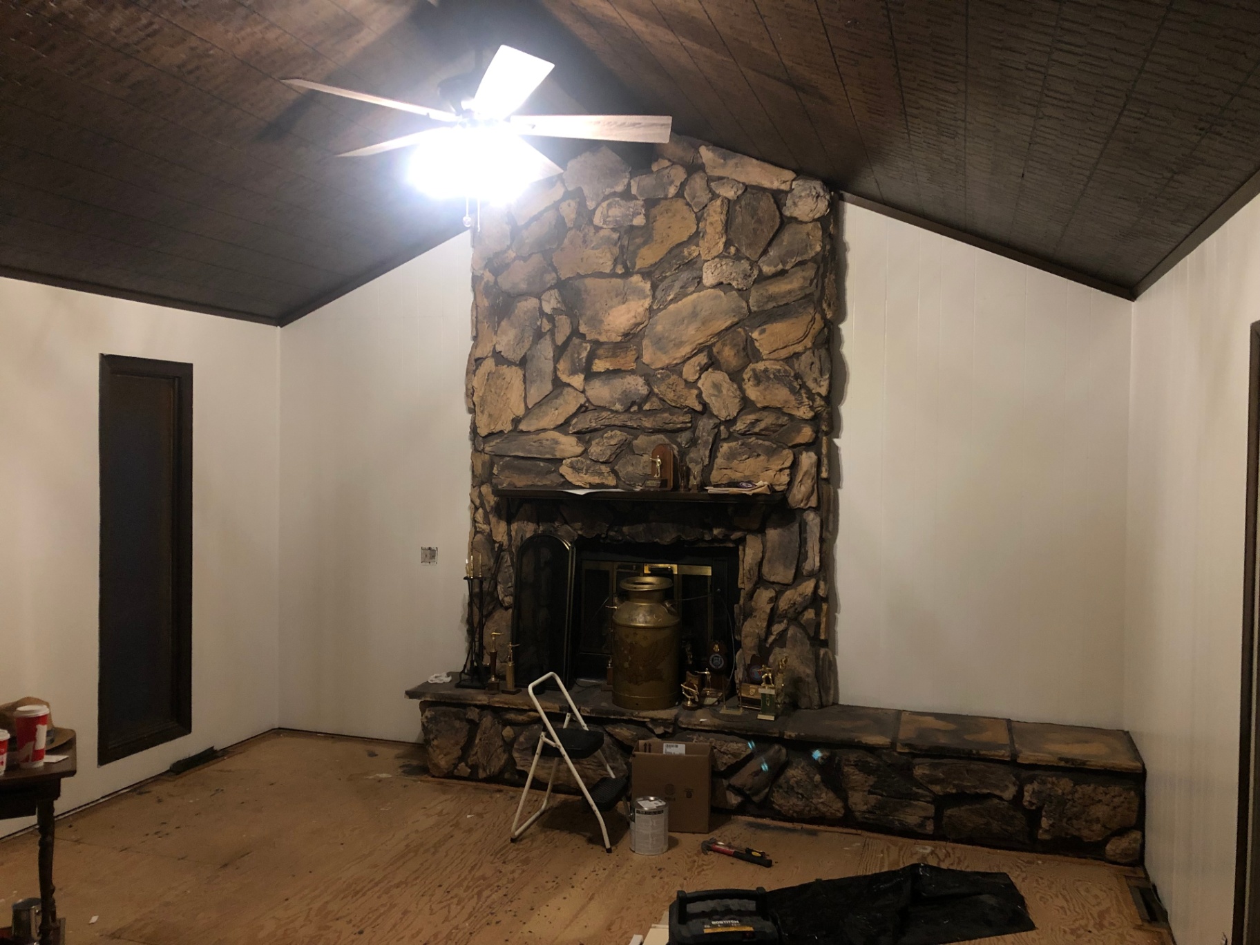 Living room with dark wood paneling repainted crisp white around the original floor-to-ceiling stone fireplace, with a dark plank ceiling and ceiling fan