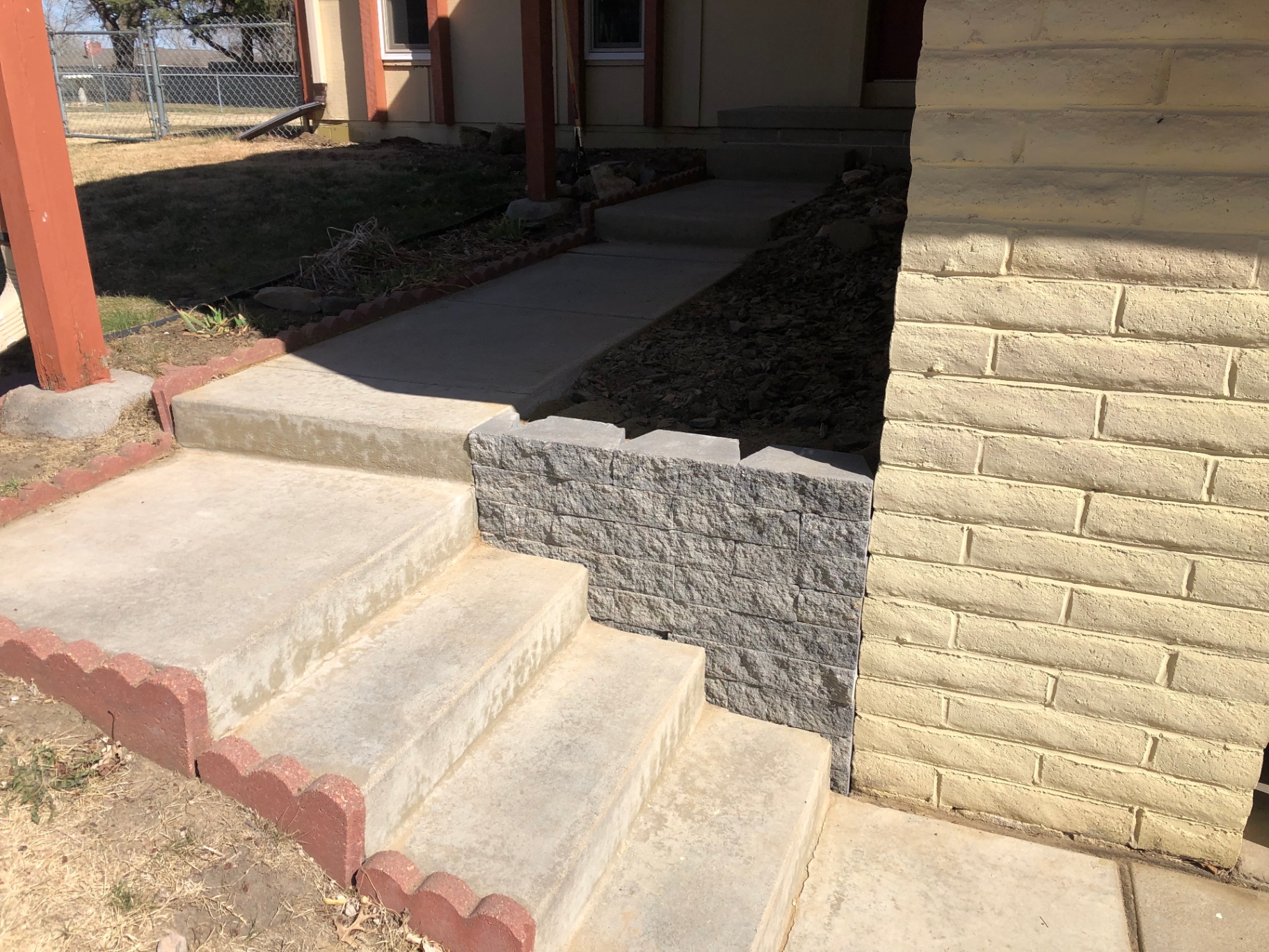 Front concrete steps with a new gray stacked-block retaining wall replacing the old loose stone stack next to a painted brick foundation